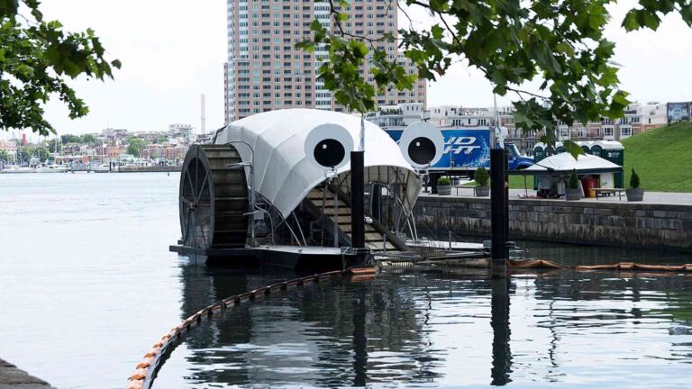 Googly eyed trash wheel hauling Baltimore harbor litter up its conveyor belt, cartoon eyes pulls